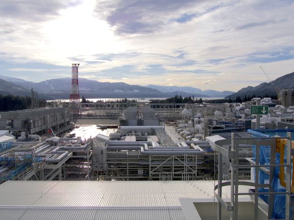 A maze of pipes and industrial machinery include a flare tower, with mountains and a ocean inlet in the background.