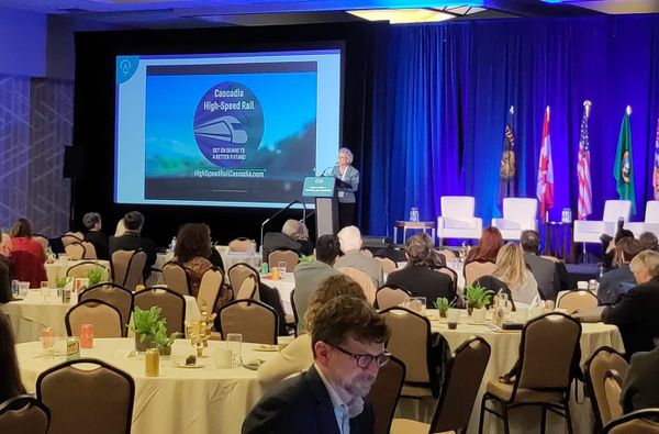 A speaker addresses a ballroom filled with various regional leaders. A power point reads Cascadia High Speed Rail. 