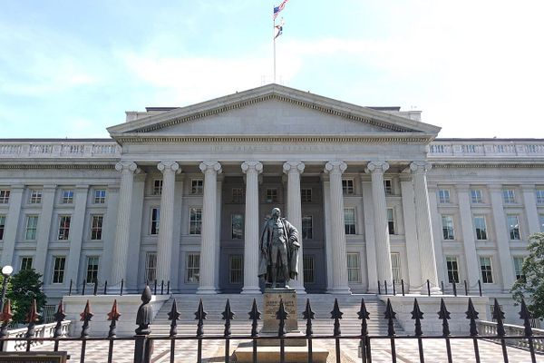 The facade of the US Treasury building with Greek columns, a statue of a revolutionary era figure and an iron gate.