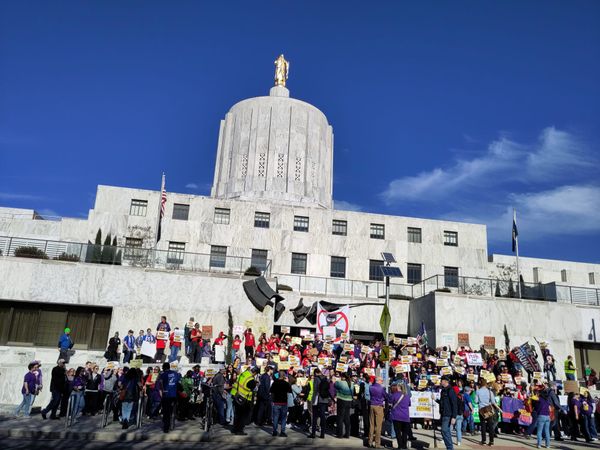 A crowd of activists with signs, including an anti-ICE sign stand in front of the Oregon state capitol building.