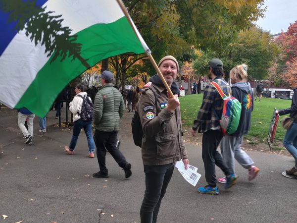 A man in a jacket with a rainbow peace patch on the shoulders holds a blue, white, and green flag of Cascadia.