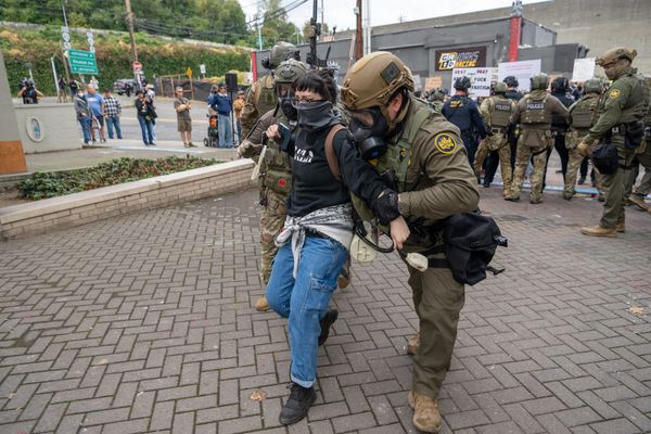 Two goons in helmets, tactical gear and gas mask hold a female protester by the arms. She wears a face mask and black hoodie.