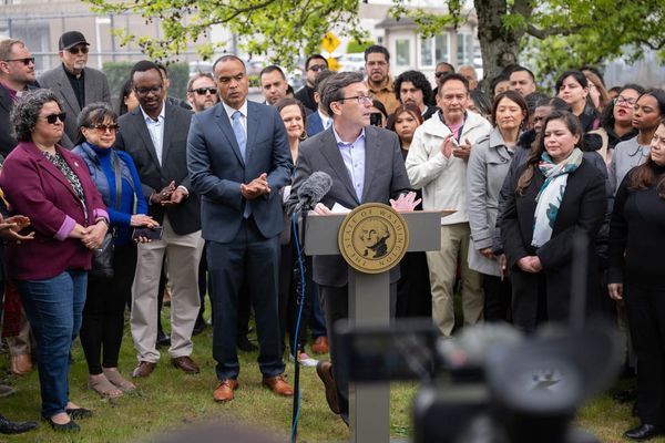 A man in a suit speaks in front of a group of activists and behind a podium with the seal of the state of Washingtons