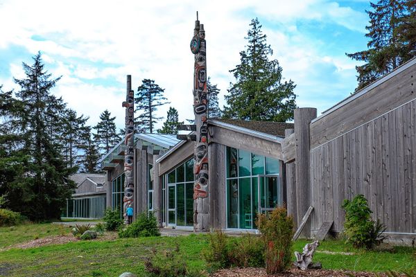A modern longhouse-inspired building with lots of glass windows, flanked by traditional Haida totems featuring figures of eagles, bears, and whales