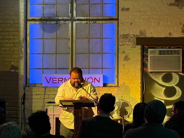 A poet reads into a microphone with a brick wall, glowing window and a sign that reads Vermillion behind him