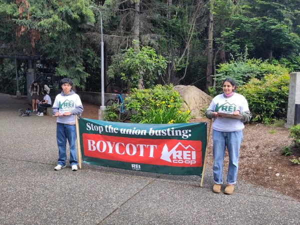 Two organizers wearing REI Union shirts hold a sign that says: Stop the union busting: boycott REI co-op in front of a forested store entrance