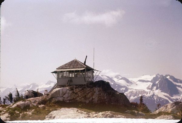 A forest lookout cabin on top of a mountain, with glaciated peaks in the distance.
