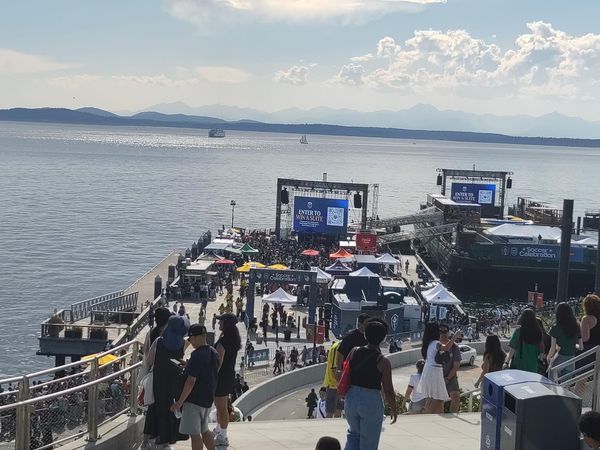 A view of a pedestrian walkway, a public TV screening area on a pier, and a vast bay with mountains and a ferry boat in the distance