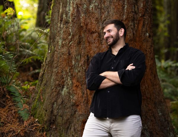 A person with a beard wearing a black shirt and tan jeans stands in front of a large evergreen tree.