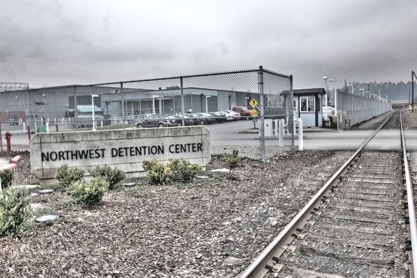 A jail behind barbed wire near train tracks and a sign that reads Northwest Detention Center.