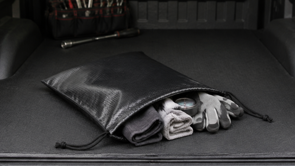 Open black PU-coated fabric drawstring bag holding work gloves, folded cleaning cloths and a round gauge in a vehicle’s cargo area.