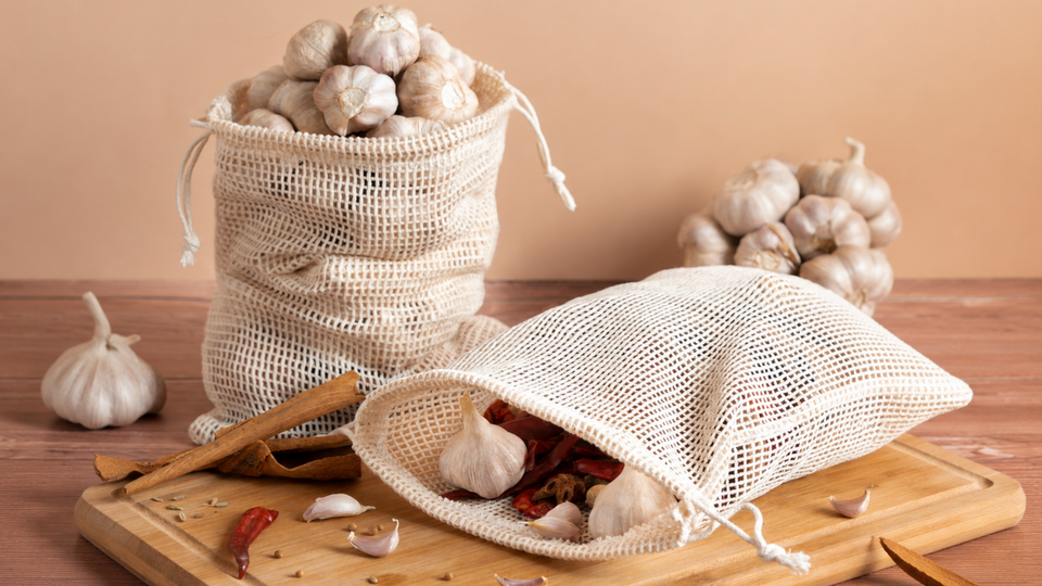 Two natural cotton mesh drawstring bags holding garlic and dried chillies on a wooden table, with cinnamon sticks and loose garlic cloves nearby.