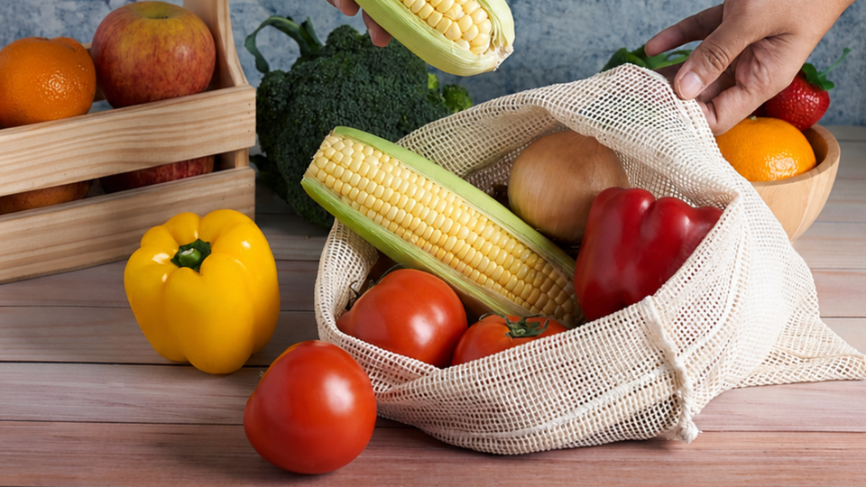 Hands placing corn into a natural cotton mesh drawstring bag filled with tomatoes, an onion and a red pepper, surrounded by fresh produce on a wooden table.
