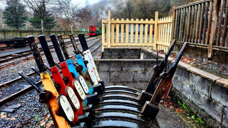 Red, yellow, blue, and white signal levers near train platform/tracks; caboose receding into the distance.