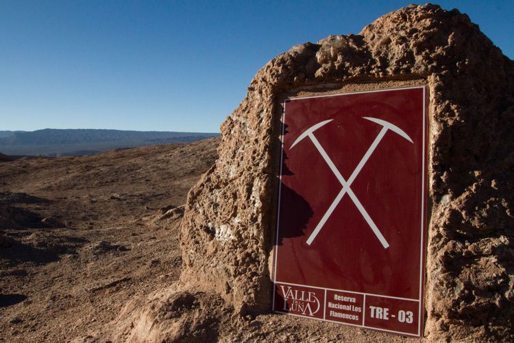 Blue sky, rolling terrain, maroon sign with grey graphic of 2 pickaxes, "Valle Dela Luna, Reserva Nacionale Los Flamencos"