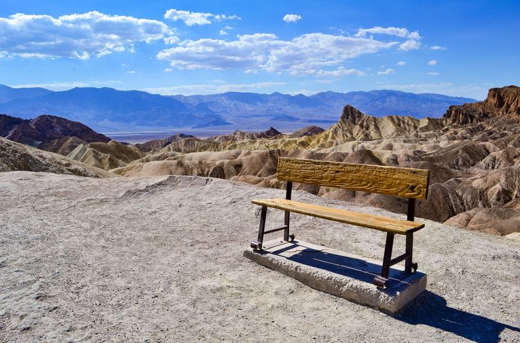 Craggy wooden bench on dusty sand, with desert mountains in the background.  Sunny day, blue sky, low clouds
