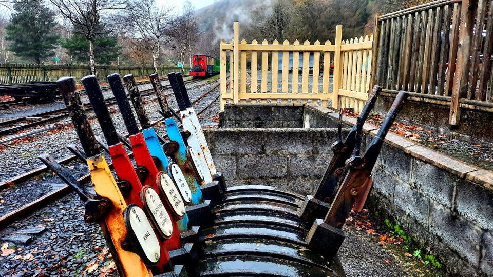 Red, yellow, blue, and white signal levers near train platform/tracks; caboose receding into the distance.