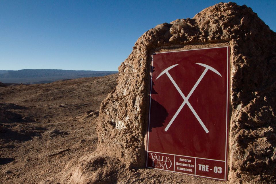 Blue sky, rolling terrain, maroon sign with grey graphic of 2 pickaxes, "Valle Dela Luna, Reserva Nacionale Los Flamencos"