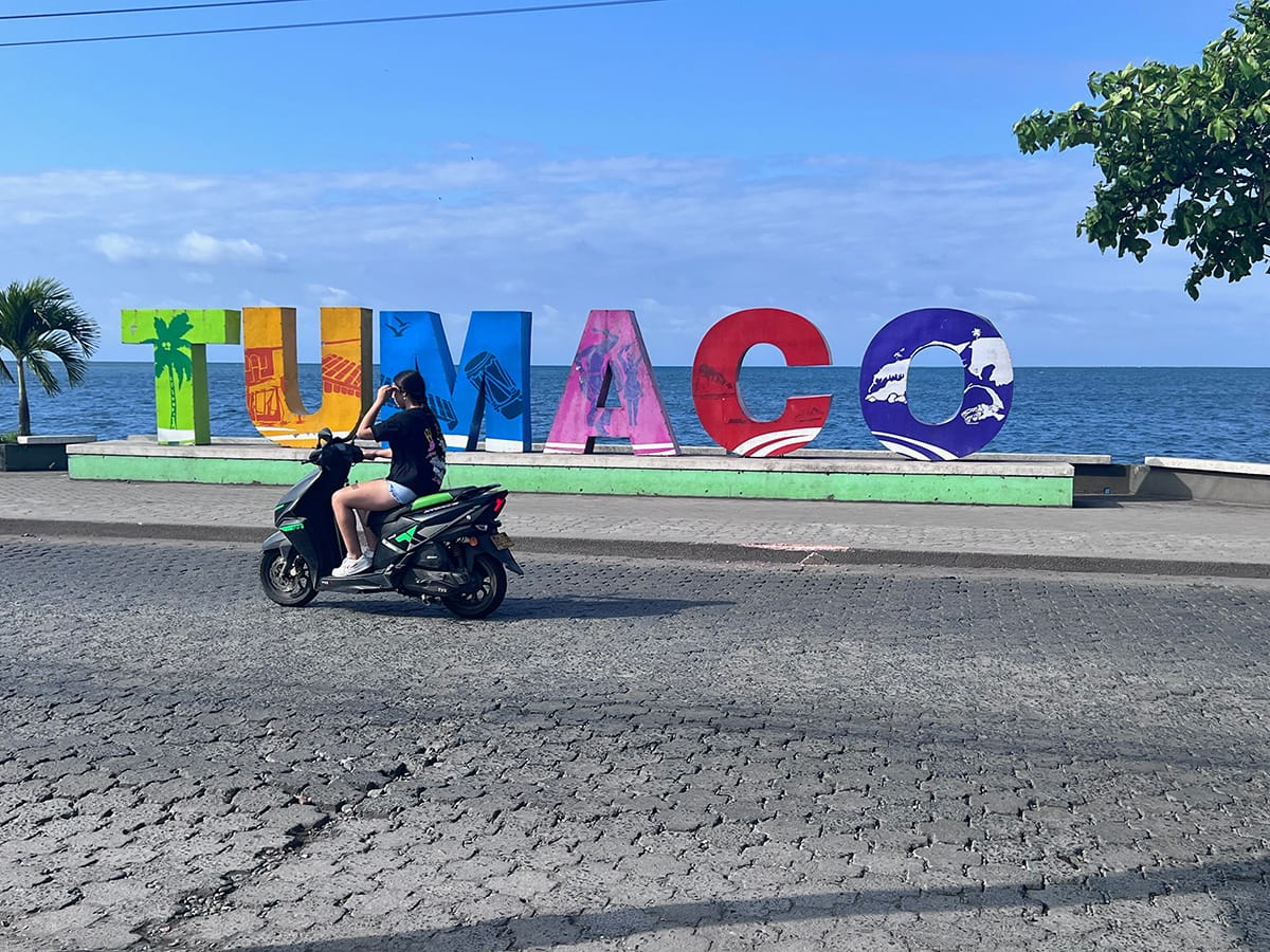 Image shows a motorcylist riding along the beach in front of a Tumaco sign