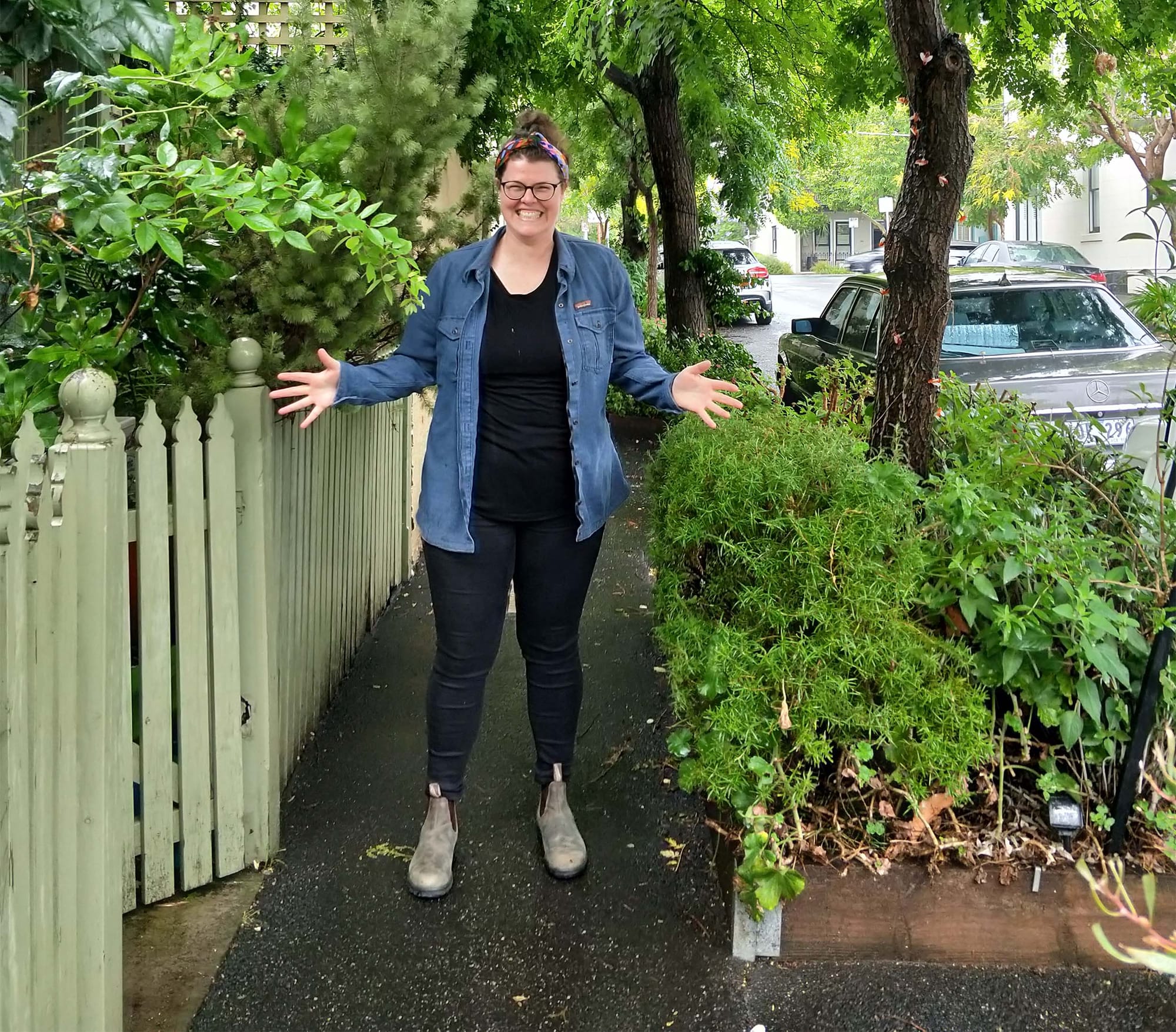 A person standing and smiling on a sidewalk, with plants and trees around them