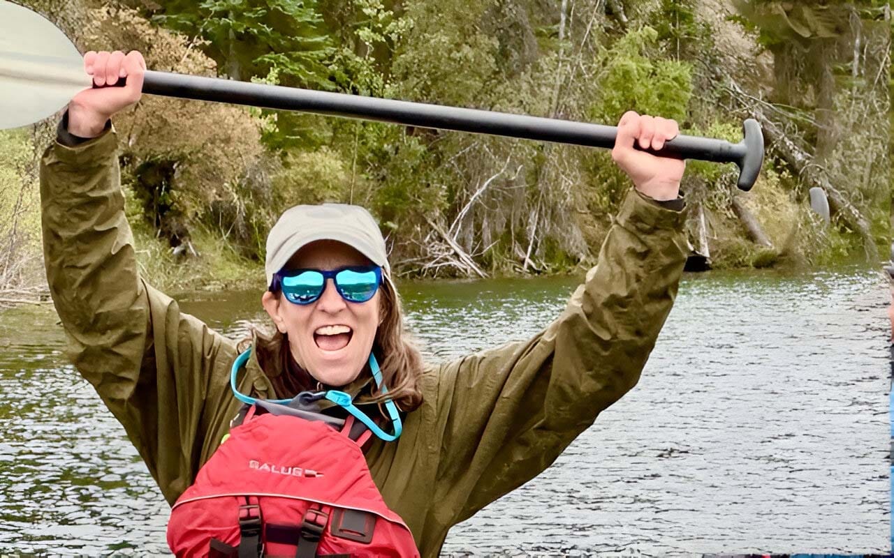 A person in sunglasses wearing a PFD, holding up a paddle with water and trees behind