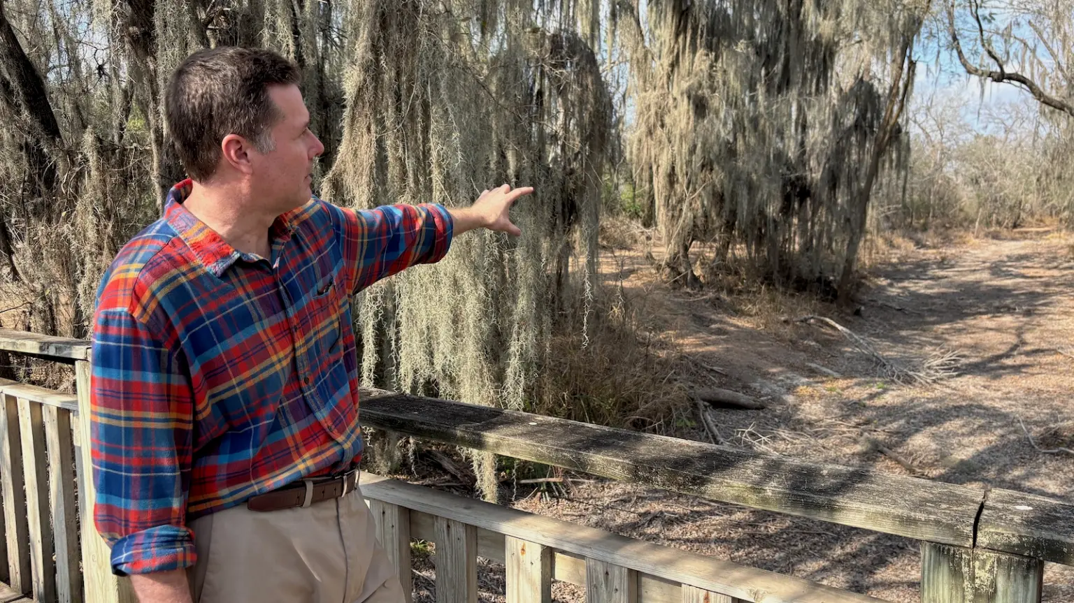 A person in a plaid shirt standing on a wooden walkway and pointing at a natural area and pathway