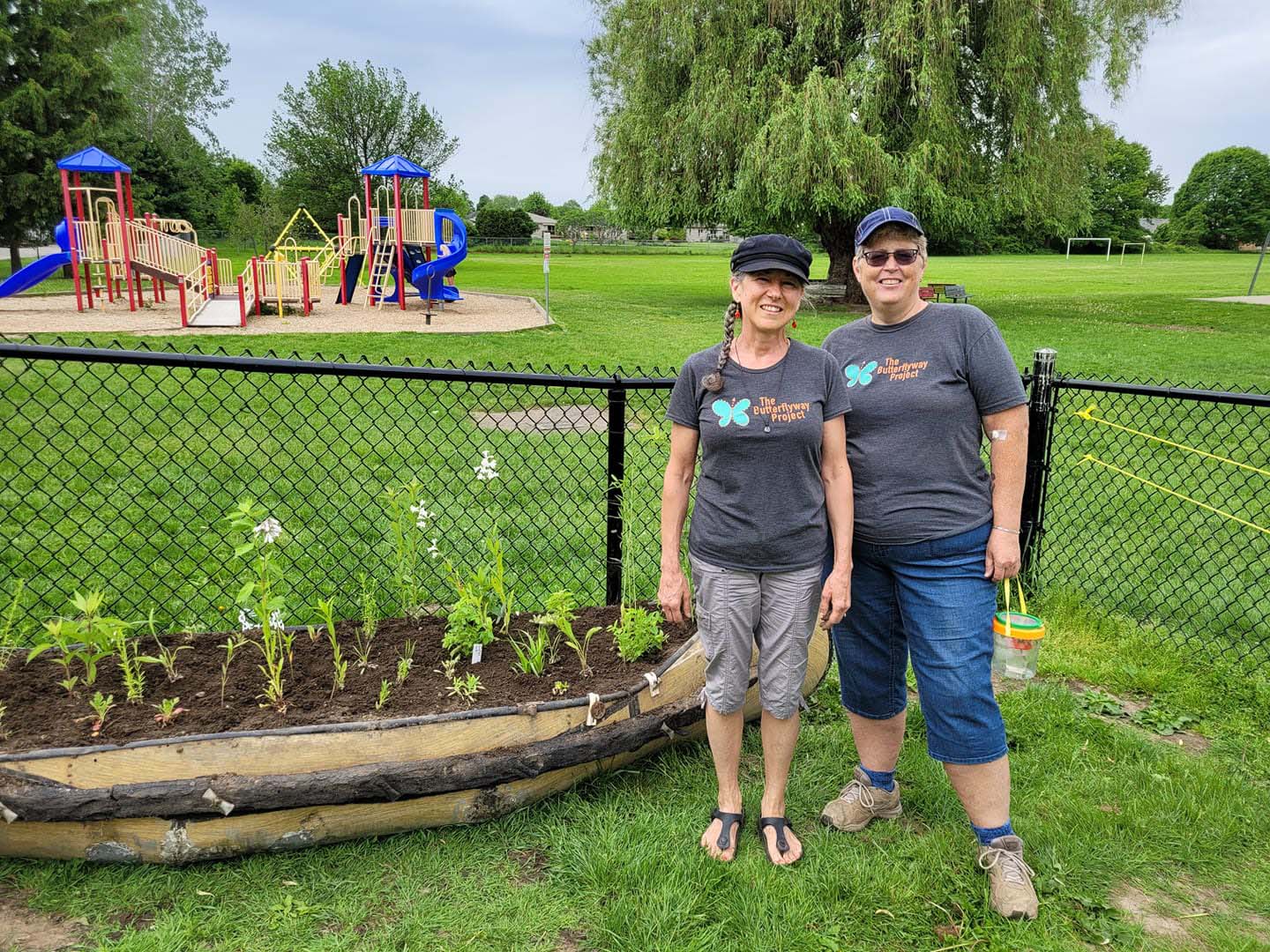 Two people posing in a grassy park next to a wooden boat holding a newly planted garden