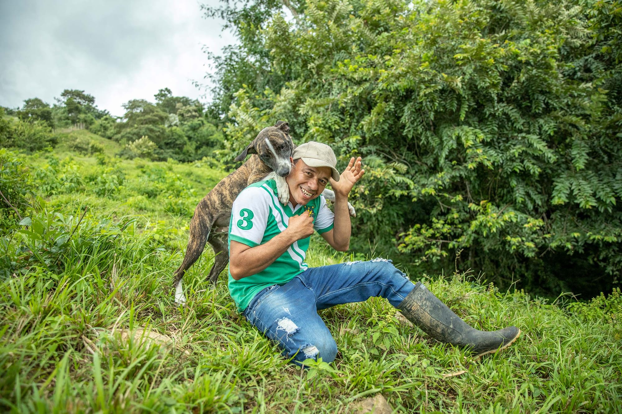 A person in jeans, an athletic shirt and rubber boots playing with a dog outdoors, amidst verdant grass and shrubs