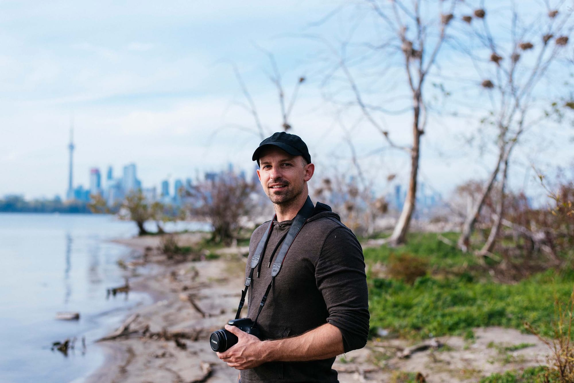 A person with a camera around is neck smiles facing the camera with a natural setting and Toronto skyline in the background