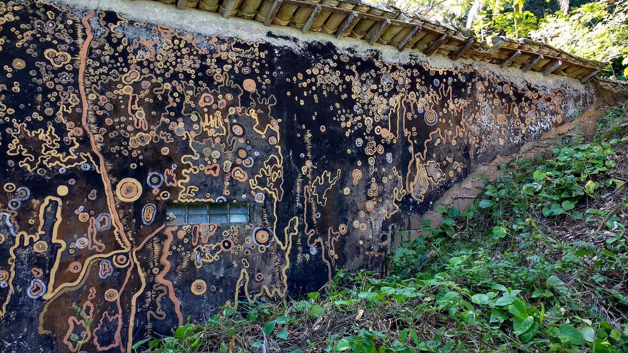The side of an old building next to a green slope, painted with circles and squiggles in earthy colours