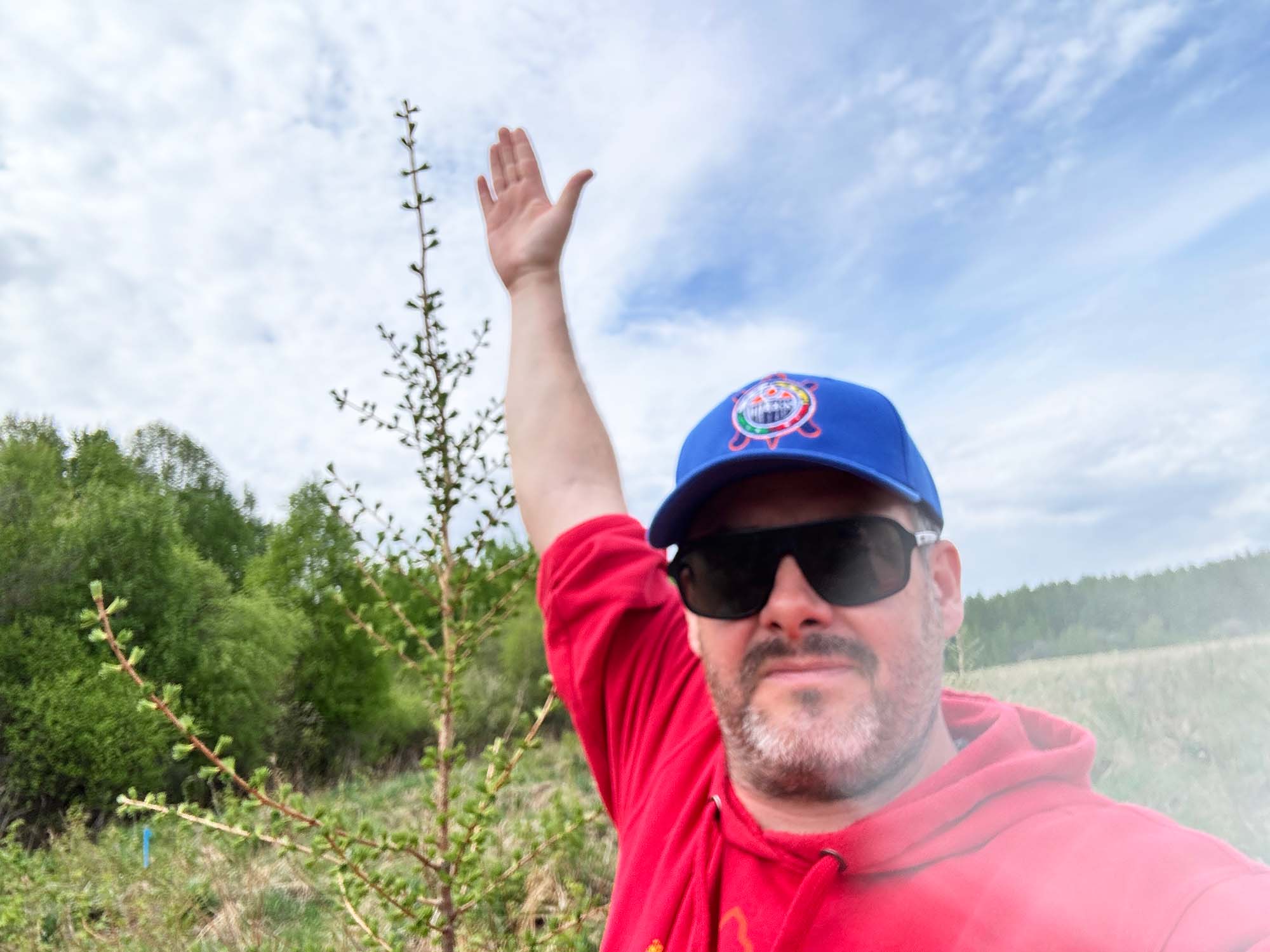 A person wearing a ball cap in an outdoor environment, gesturing at the height of a young tree