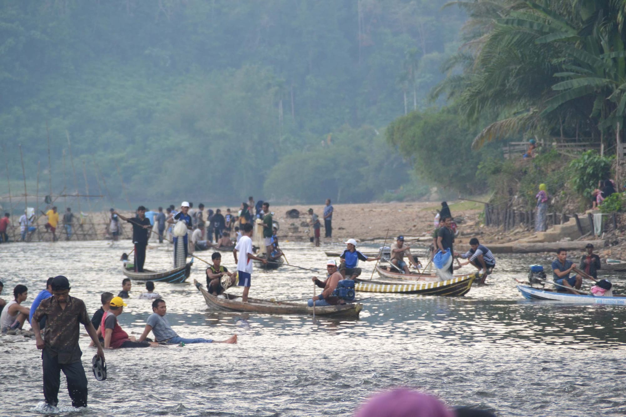 People and small boats in a river with forested hillside beyond