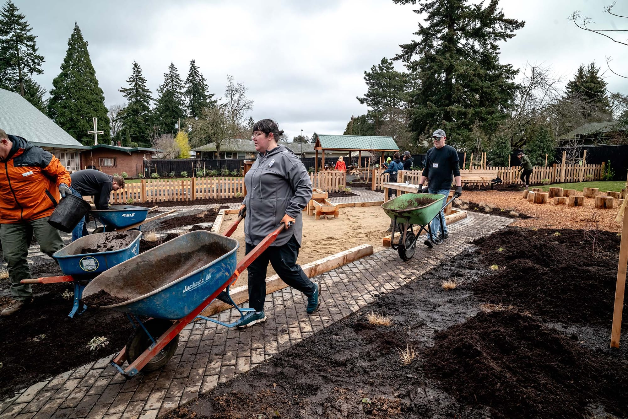 People pushing wheelbarrows across a pathway through a landscaped outdoor space