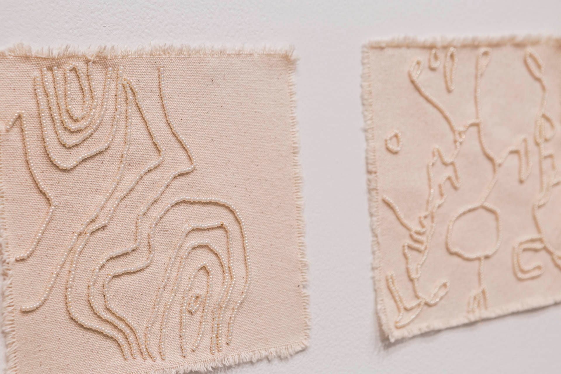 Two squares of cream-coloured cloth hanging on a white wall, each stitched with curved lines of cream-coloured beads