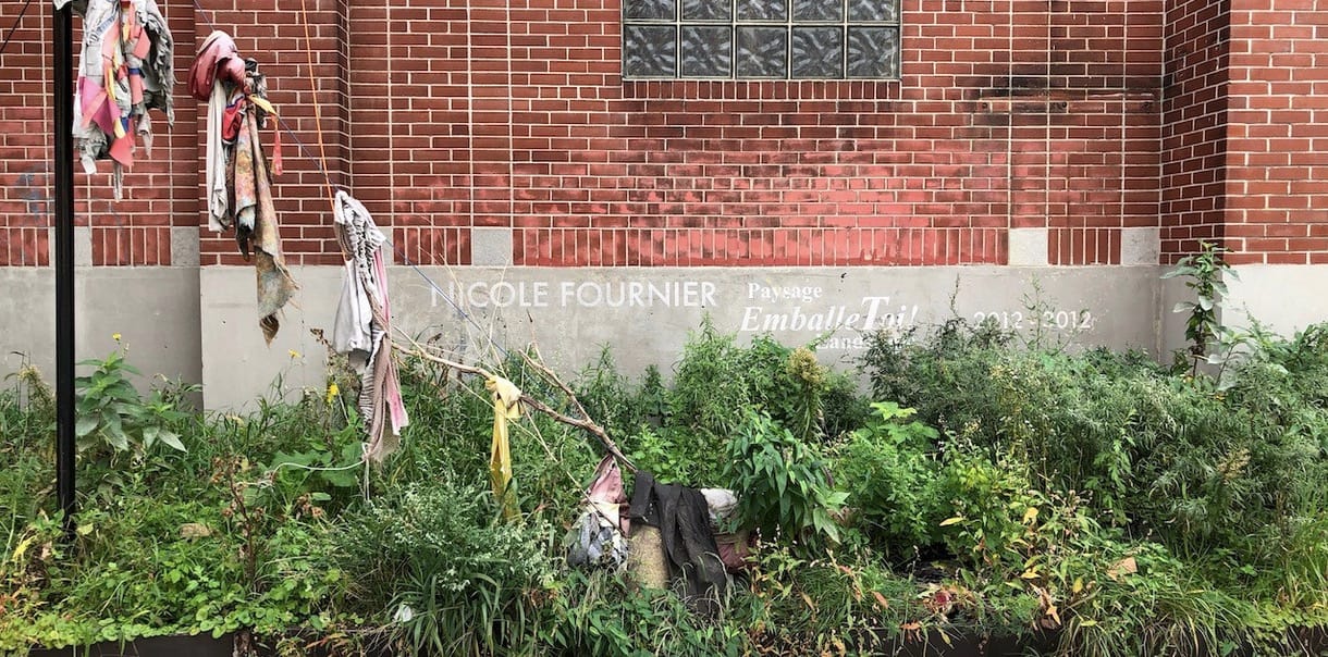 Fabric strung and tied on wire and branches in front of a red brick wall and above thick low greenery
