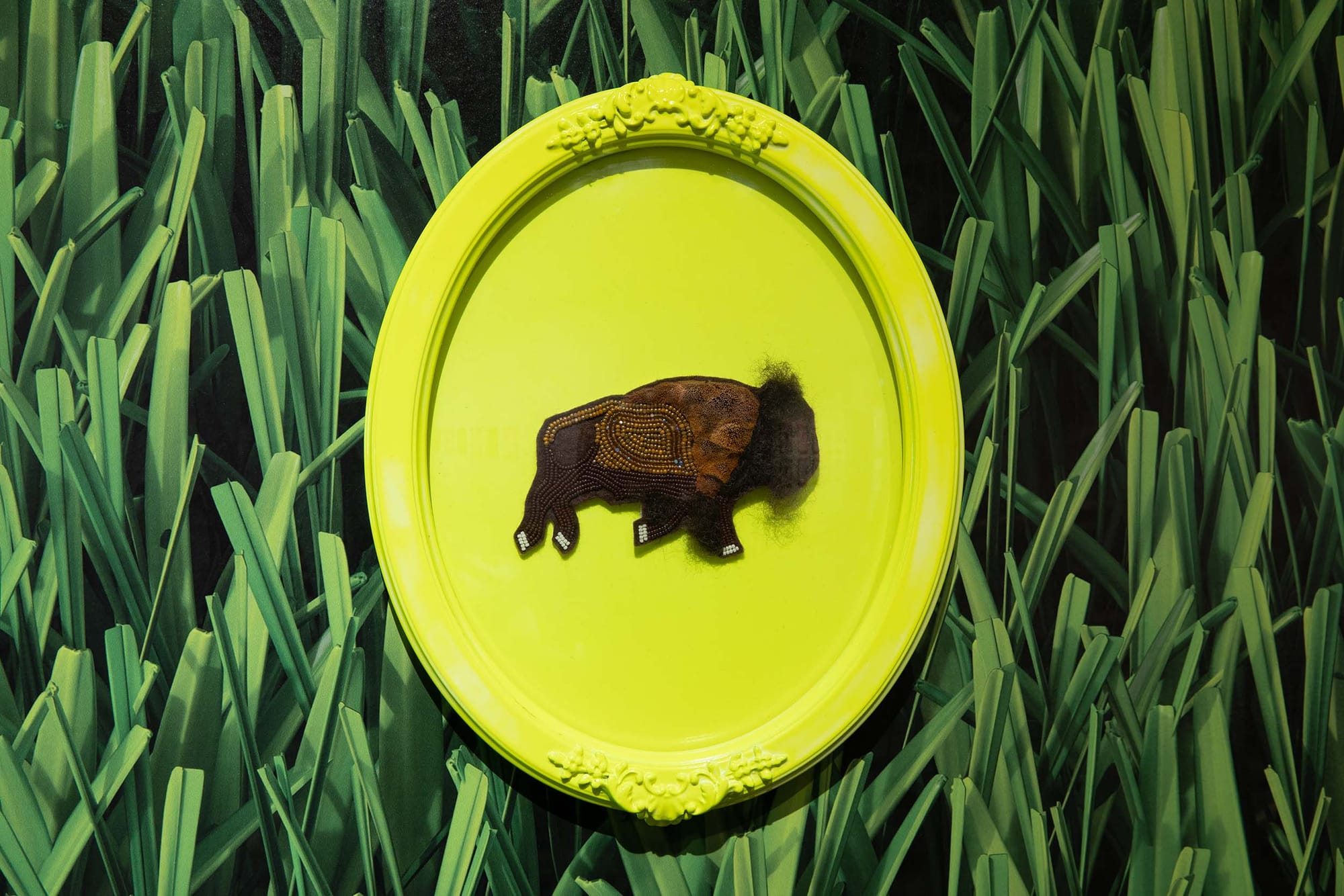 A sewn and beaded bison art piece on a bright green oval frame with an image of grass behind