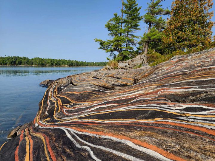 A lakeside rocky outcrop painted with earthy mineral stripes, on a sunny blue-sky day