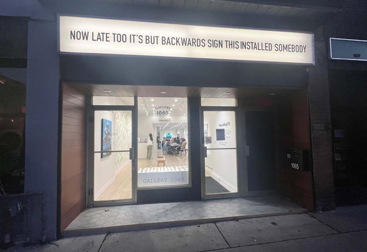 Front view of a small gallery from the sidewalk. Indoors, lights are on and people sitting around a table.