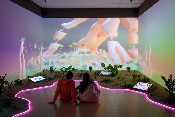 Two people taking in a multimedia art exhibition featuring digital screens, pink lighting and plants