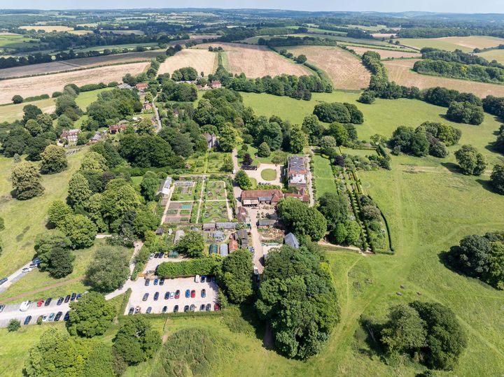 Aerial view of land with buildings, gardens and a car park in the centre surrounded by trees and fields