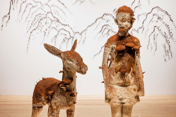Close-up of art pieces: a small deer and a person in brownish white with mushrooms emerging, standing on hardwood floor with wall behind