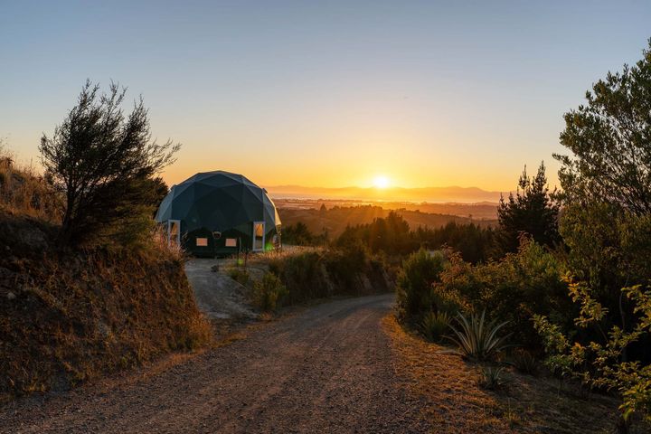 A geodesic dome next to a gravel road amidst brush, with blue sky and sunset beyond.