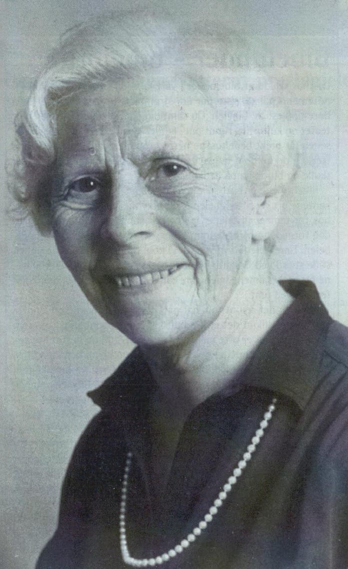 Portrett av Elna Kvellestad, en eldre kvinne med hvitt hår og perlekjede, som smiler