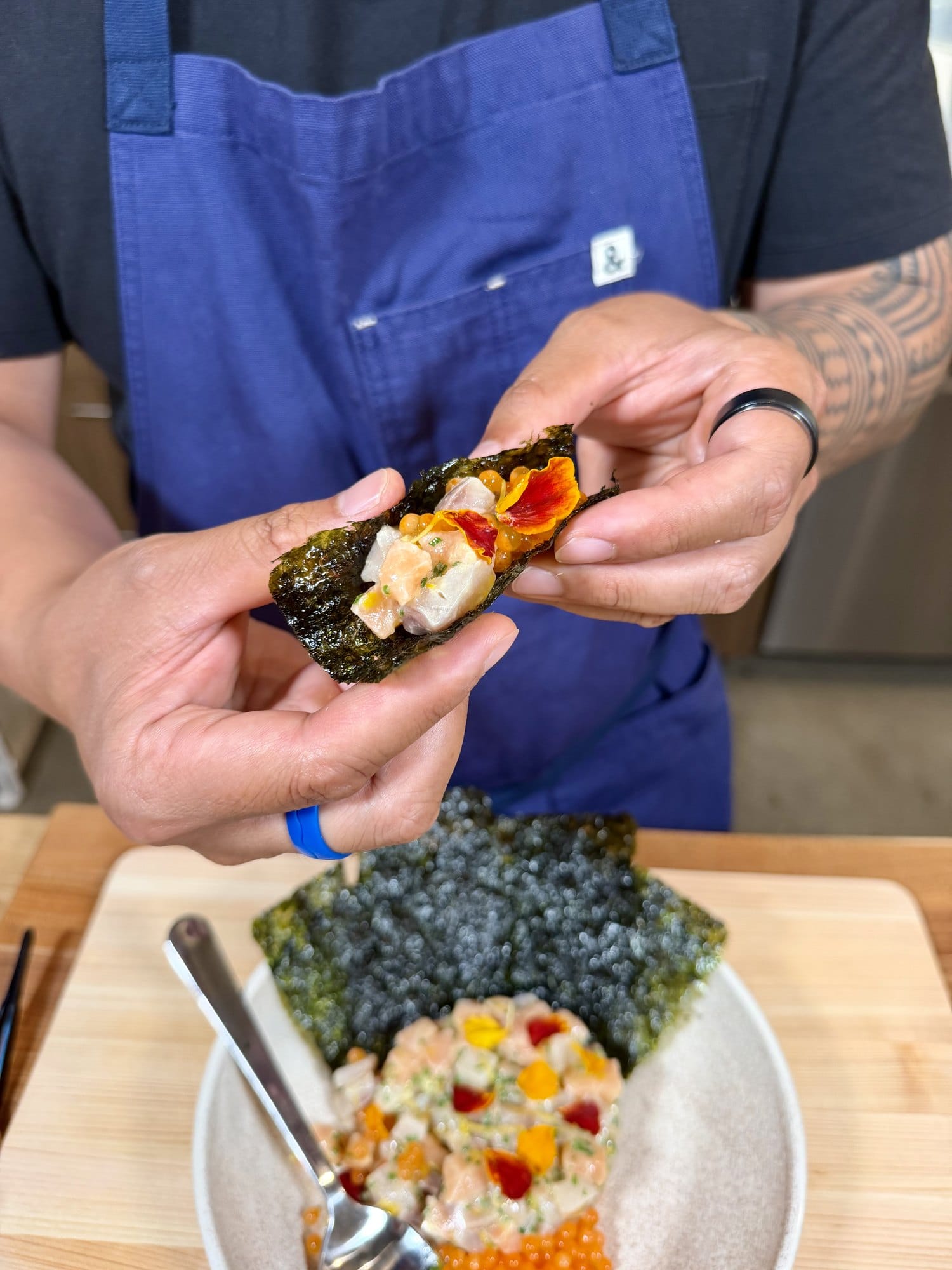 A hand holding toasted nori wrapped around tartare and salmon roe above the plated dish.