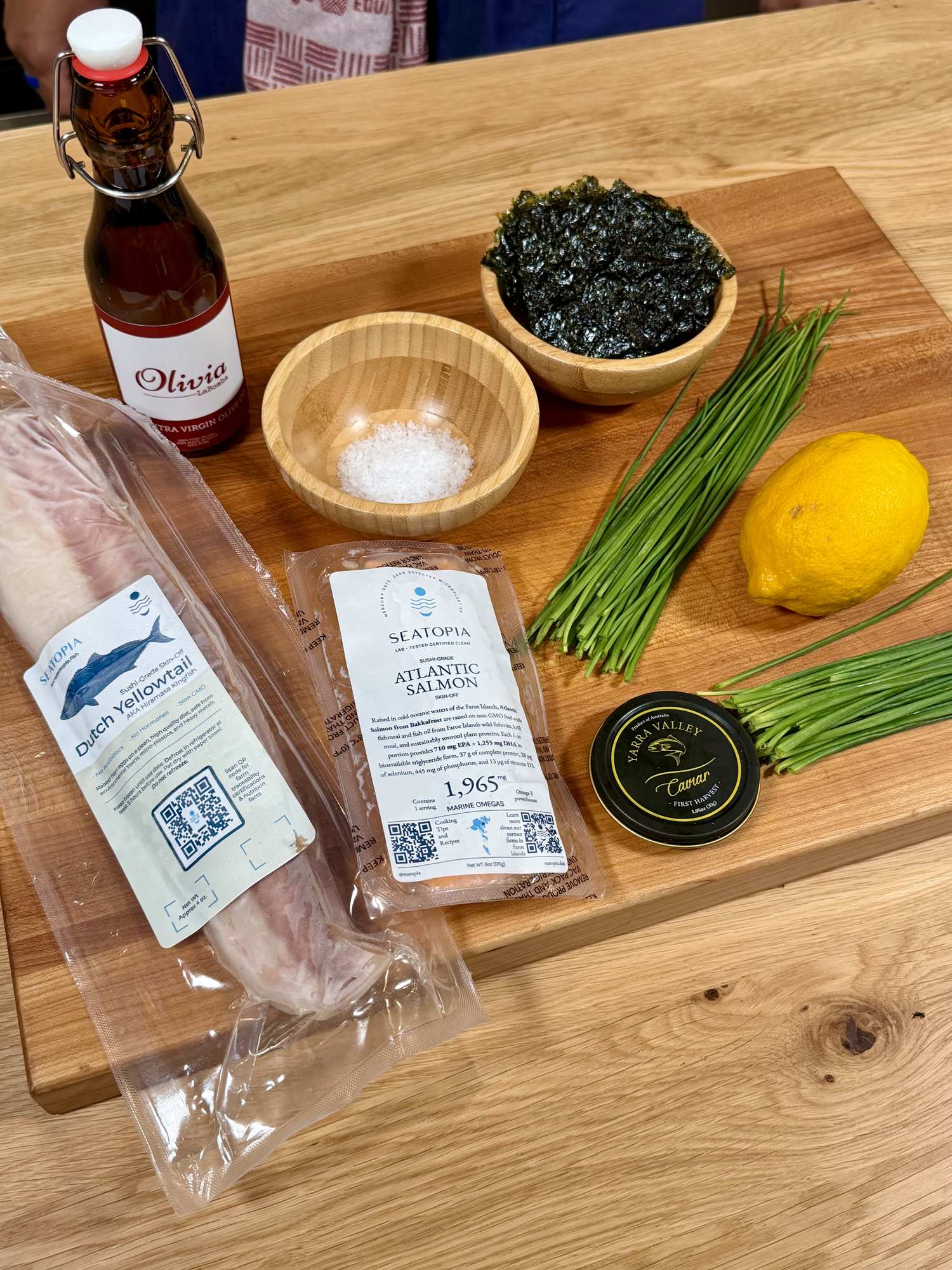 Tartare ingredients on a board: olive oil, flaky salt, nori, chives, lemon, sealed fish, salmon roe.
