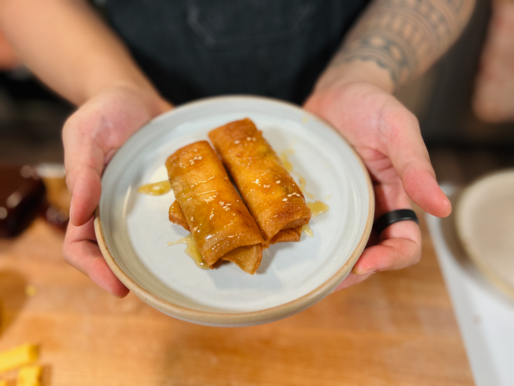 Golden-brown lumpia drizzled with honey and sesame seeds on a white plate, held by hands