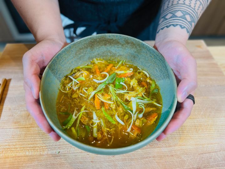 Hands holding a turquoise ceramic bowl of golden-orange chicken sotanghon soup with translucent noodles, sliced carrots, and green scallions garnish on a wooden cutting board.