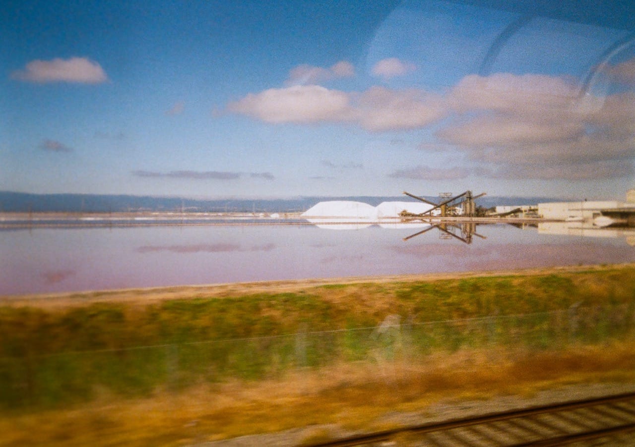 a color photo of some agricultural equipment in the distance, reflected in a pond, under a blue sky dotted with clouds. blurry train tracks in the foreground
