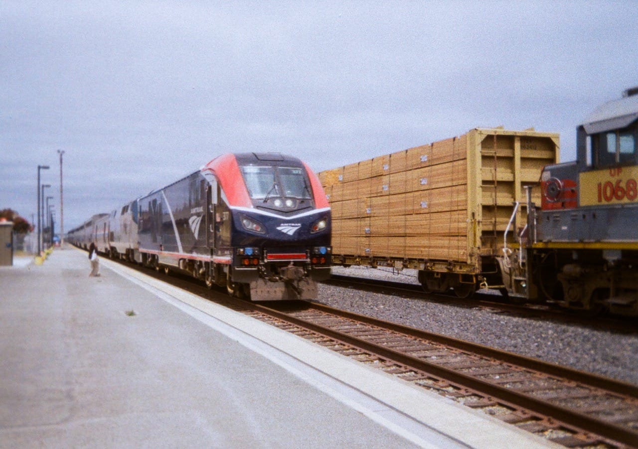a color photo of Amtrak's Coast Starlight train approaching Salinas Station alongside a freight train hauling lumber