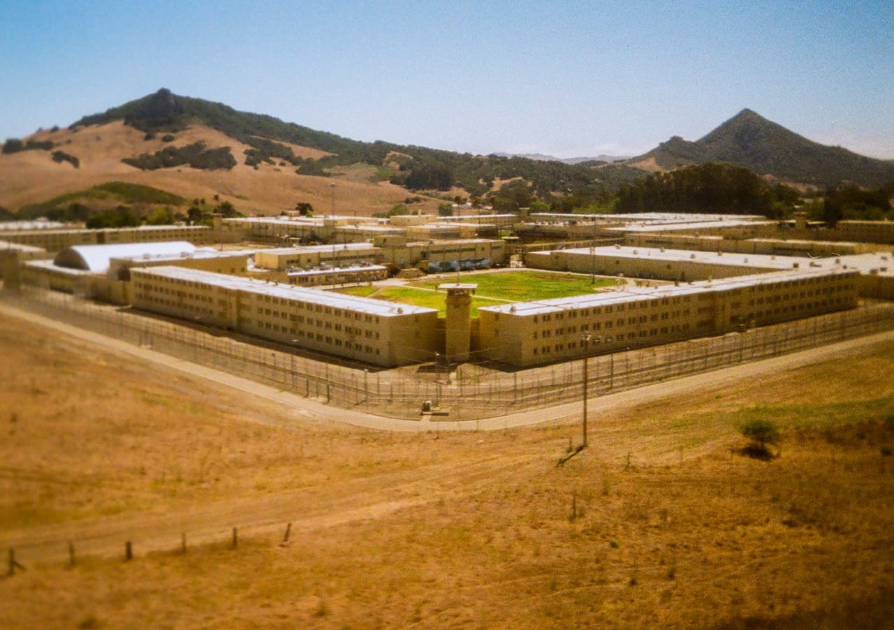 a color photo of a prison complex behind security fencing. brown grass in the foreground, and two mountain peaks on either side in the background